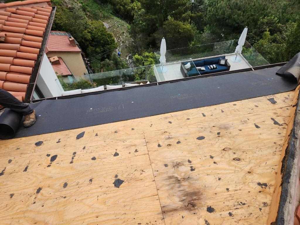 Bare wood roof deck next to a dark strip of underlayment and an adjacent tiled slope during roof work